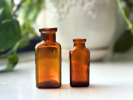 Two small, amber-colored glass bottles standing side by side on a white surface. The bottles are rectangular in shape and have a slightly flared base. The larger bottle is on the left and the smaller bottle is on the right.