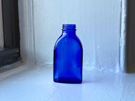 Vintage cobalt blue glass medicine bottle standing on a white windowsill, illuminated by soft natural light, showcasing its vibrant color and rectangular shape – Brooklyn Sea Glass