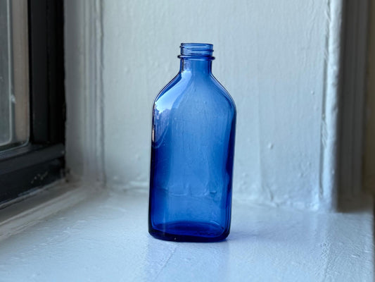 Vintage cobalt blue Phillips Milk of Magnesia bottle standing upright on a white windowsill reflecting its rich blue hue – Brooklyn Sea Glass