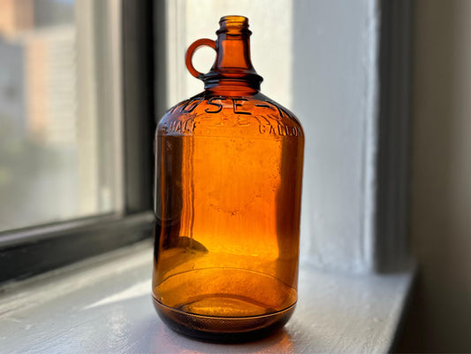 Vintage amber glass bottle with a carrying handle. The bottle has raised lettering that says ROSE-X and ONE GALLON.