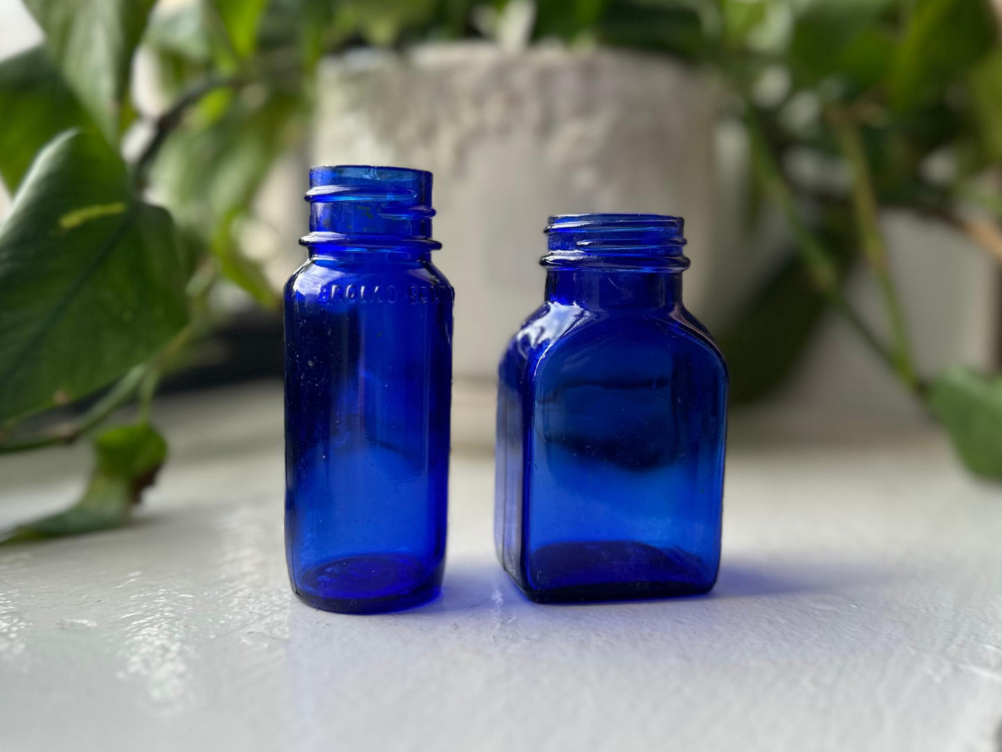 Two small, blue glass bottles standing side-by-side on a white surface. The bottles are different shapes and sizes, with one being rectangular and the other being more rounded. They are both slightly tilted and have a plant in the background.