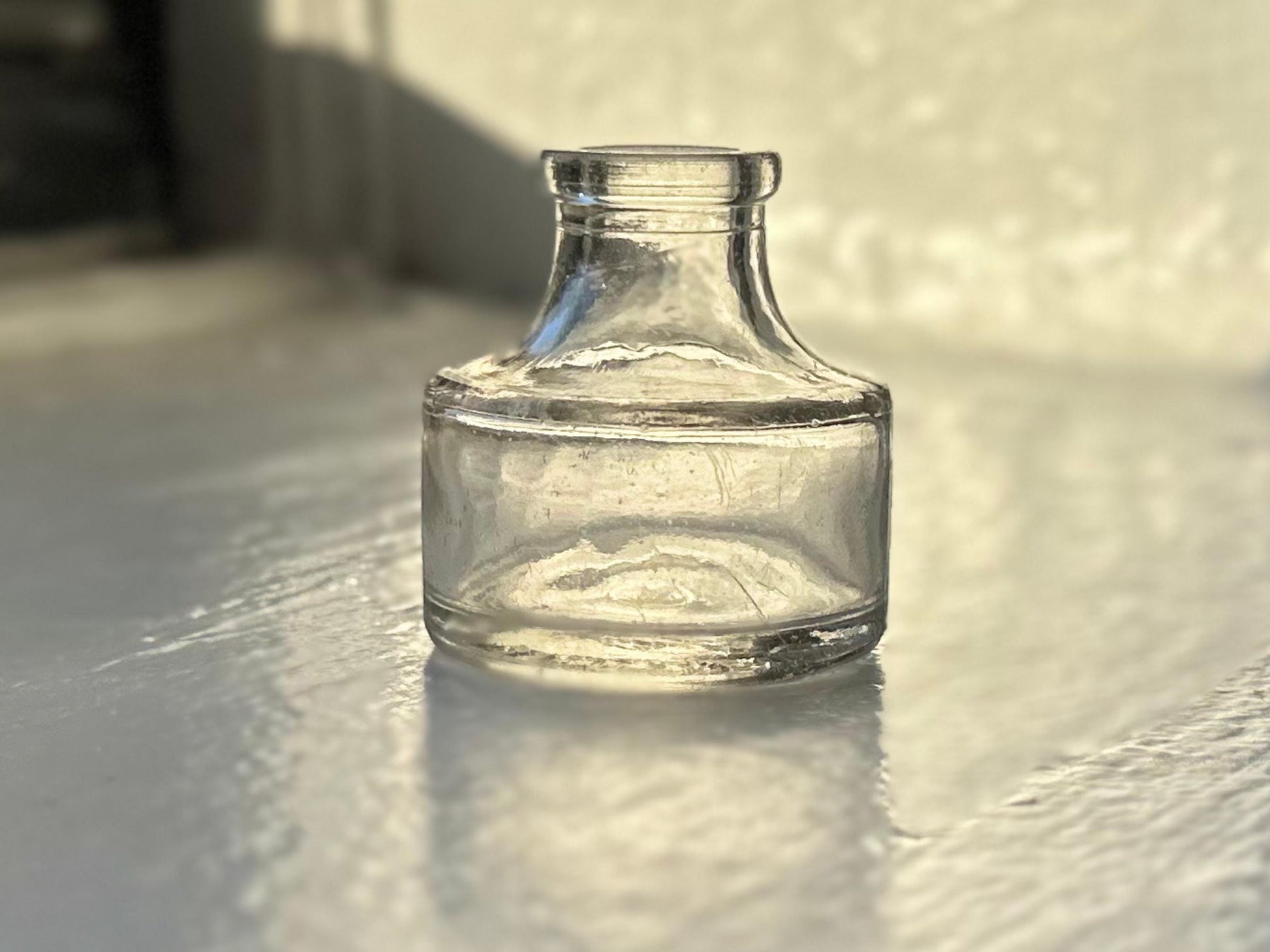 A small, antique Higgins Ink Jar from 1947. The jar is clear glass with a cylindrical shape and a short neck. It sits on a white surface with a slight reflection.
