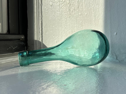 A vintage teal glass bottle with a bulbous shape and a narrow neck, resting on a white surface. The bottle has a slight greenish tint and appears to have been hand-blown.