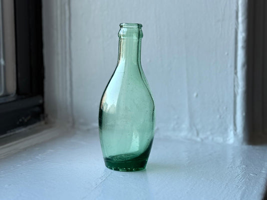A green vintage Perrier glass bottle standing on a white windowsill with soft natural lighting, highlighting its subtle curves and embossed base.