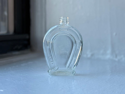 Front view of a vintage Gourelli horseshoe-shaped perfume bottle in clear glass, displayed on a white windowsill with soft natural light.