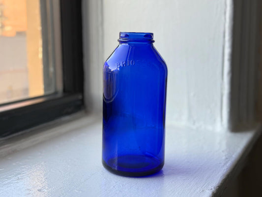 Side view of a cobalt blue Brioschi glass bottle, sitting on a windowsill with natural light showcasing its rich color and embossed branding.