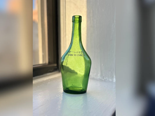 A vintage green glass Sterzi bottle standing on a white windowsill, showcasing its unique curvy shape and embossed Italian branding.