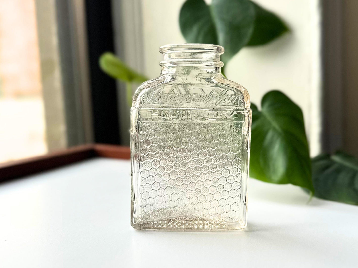 Front-lit portrait of a vintage Lake Shore Honey jar against soft natural light. The embossed “Lake Shore HONEY” text and honeycomb texture are clearly visible, with the glass’s slight pink hue enhanced by backlight. The upright stance emphasizes its clean lines and mid-century shape—an ideal accent for fall decor, farmhouse shelves, or eco-friendly displays with antique charm.