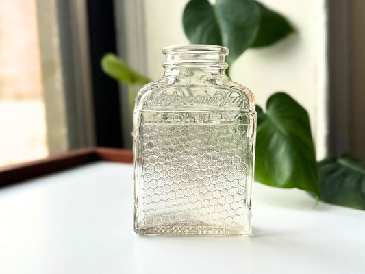 Front-lit portrait of a vintage Lake Shore Honey jar against soft natural light. The embossed “Lake Shore HONEY” text and honeycomb texture are clearly visible, with the glass’s slight pink hue enhanced by backlight. The upright stance emphasizes its clean lines and mid-century shape—an ideal accent for fall decor, farmhouse shelves, or eco-friendly displays with antique charm.