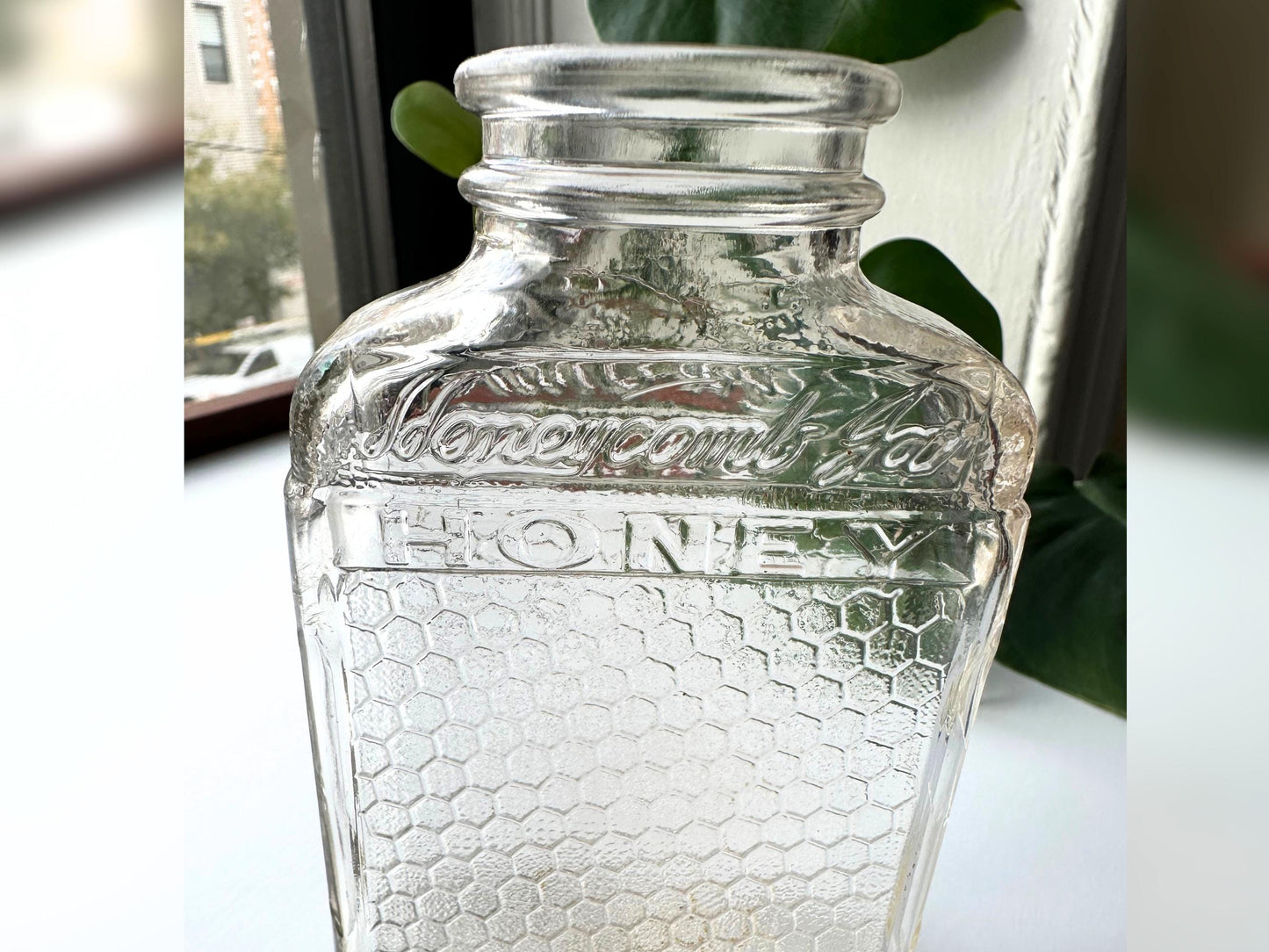 This image captures a close-up of the embossed text on the front shoulder of the vintage glass Lake Shore Honeycomb jar. The clear glass with a soft pink hue features the words “Honeycomb Jar” and “HONEY” in raised letters above a pattern of pressed hexagonal cells resembling a beehive. The intricate detailing, combined with the sturdy, thick glass construction, evokes a mid-century charm, making it perfect for rustic or sustainable decor settings.