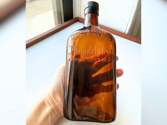 Hand holding vintage 1950 Philadelphia whiskey bottle showing true size scale and warm amber glass color in natural light, authentic post-prohibition era liquor bottle perfect for bar cart styling, vintage spirits collection, rustic home bar decor, antique bottle displays, sustainable rescued treasure
