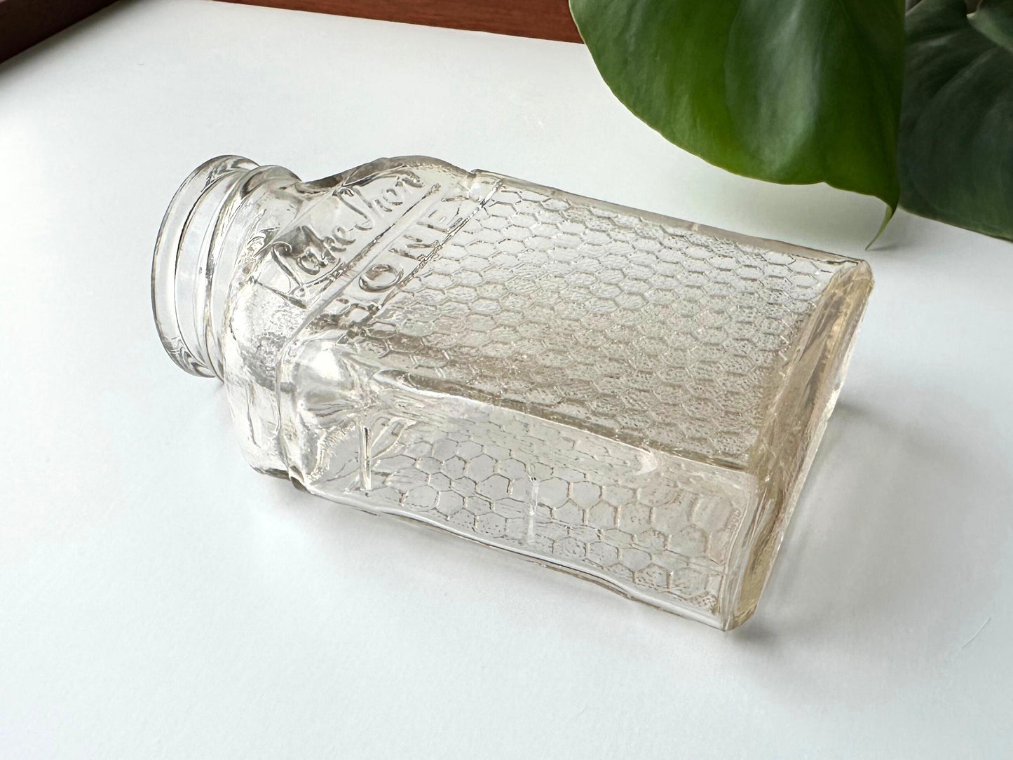 An angled side view of a vintage 1950s Lake Shore honey jar, emphasizing the honeycomb-textured glass surface and the curvature of the bottle’s shoulders. The jar rests on its side, displaying its rectangular base and highlighting its depth. Its embossed “Lake Shore HONEY” branding and apothecary style evoke a rustic charm suited to vintage farmhouse kitchens, fall-inspired table settings, or mid-century display shelves. A sustainable choice for decor or reuse in the home.
