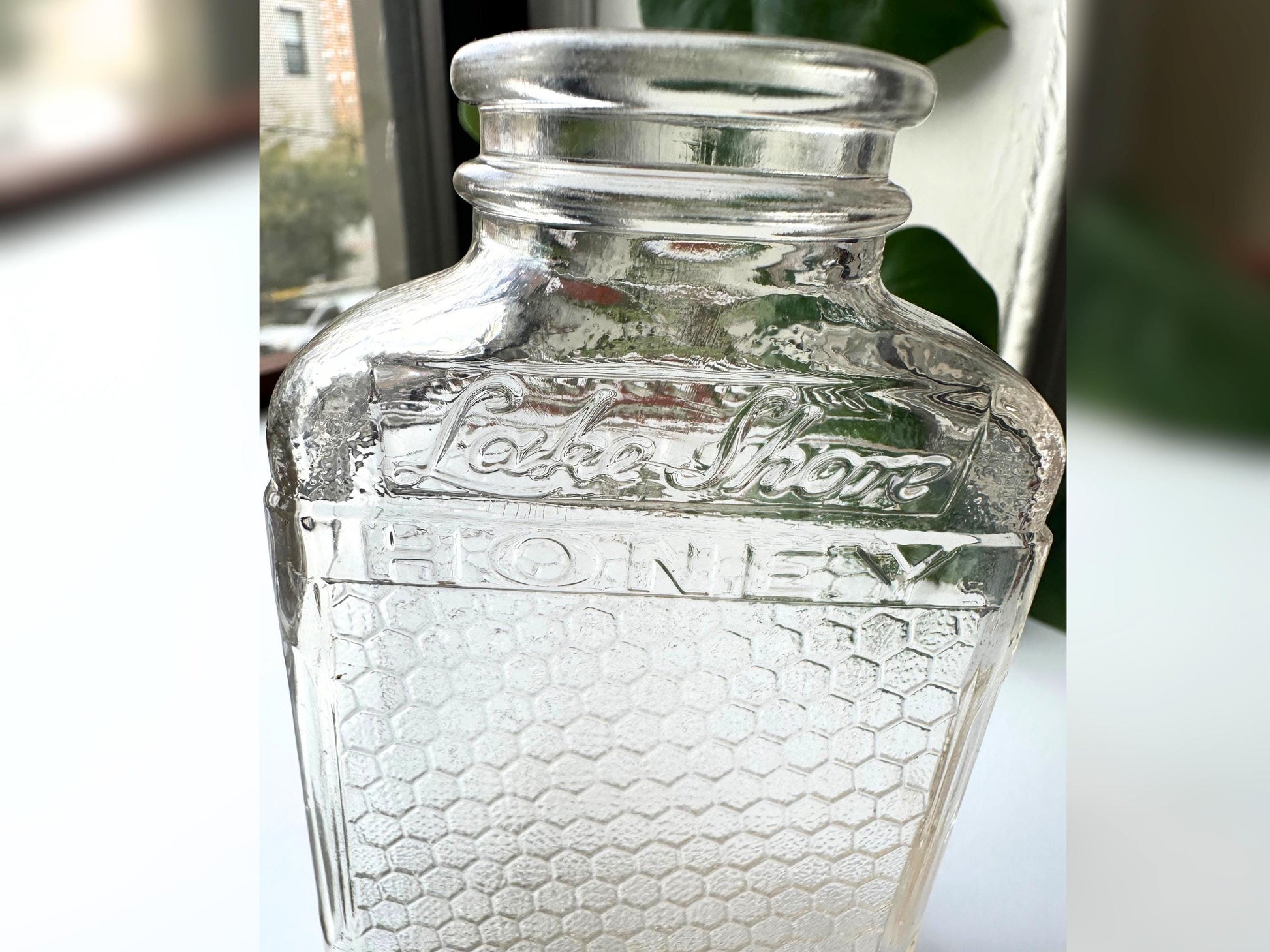 Close-up shot of the embossed “Lake Shore HONEY” text on the front panel of a vintage glass honeycomb jar. The stylized script is surrounded by hexagonal patterning, highlighting the craftsmanship and texture. The glass shows gentle wear, adding character. Perfect for detail-focused shoppers, collectors, or stylists seeking mid-century packaging design and sustainable, decorative glass.