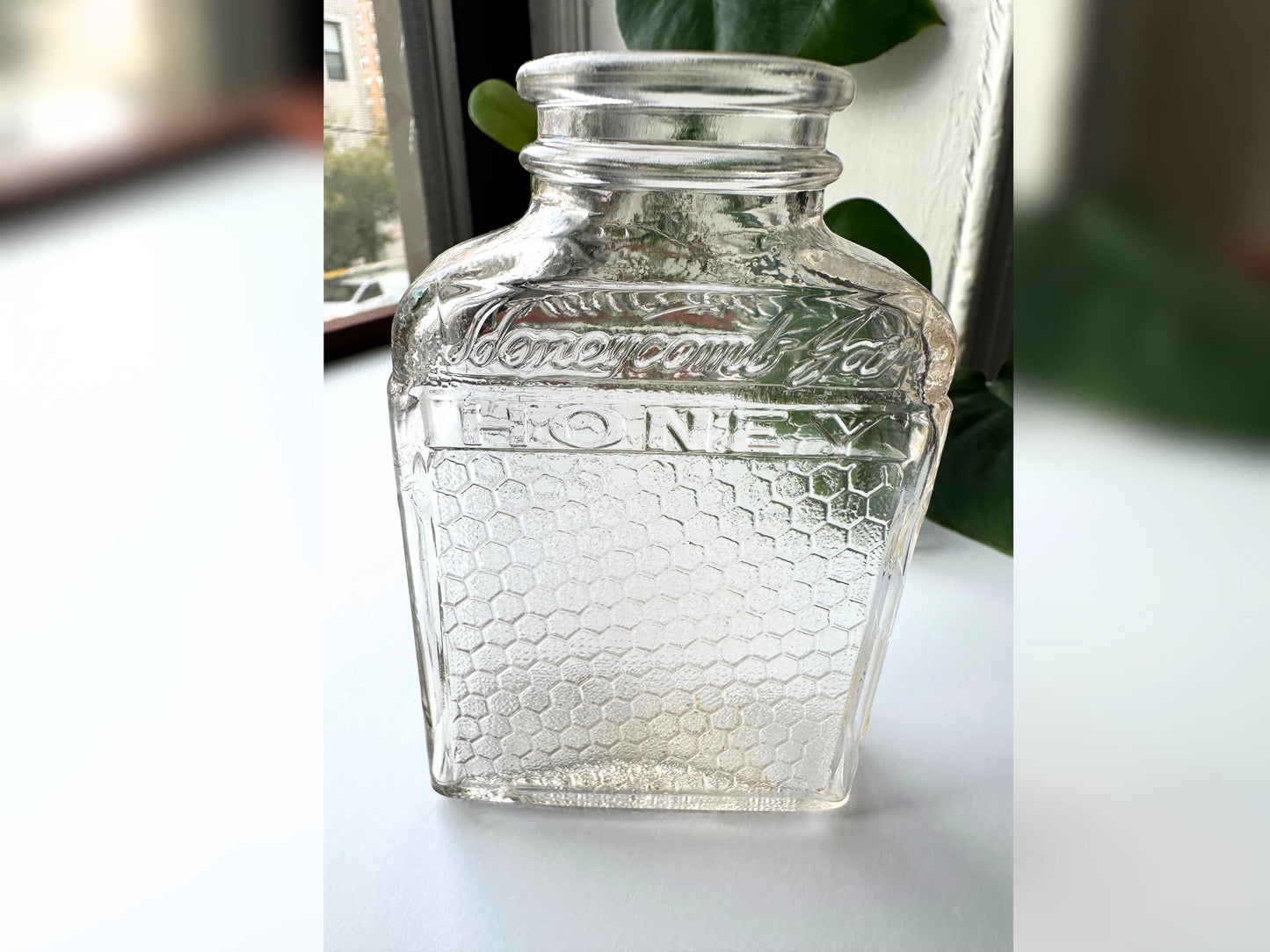 Window-lit photo of the vintage Lake Shore Honey jar upright in a moody, natural-light setting. The pink-tinted glass softly glows, highlighting the embossed branding and honeycomb design. Styled as a standalone accent, this image evokes cozy fall interiors and highlights the jar’s ability to blend into reclaimed, upcycled, or rustic vintage themes with authenticity and texture.