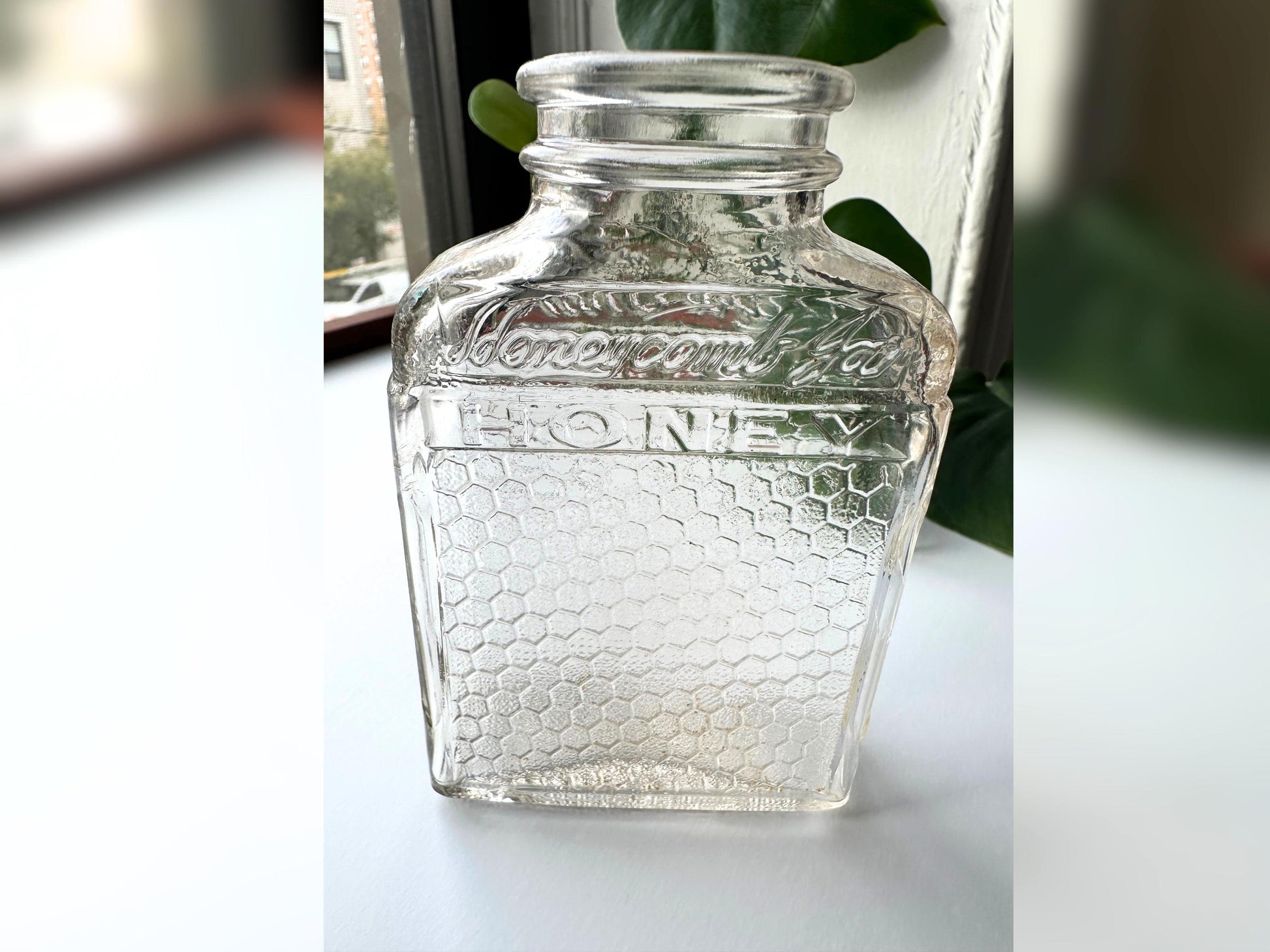 Window-lit photo of the vintage Lake Shore Honey jar upright in a moody, natural-light setting. The pink-tinted glass softly glows, highlighting the embossed branding and honeycomb design. Styled as a standalone accent, this image evokes cozy fall interiors and highlights the jar’s ability to blend into reclaimed, upcycled, or rustic vintage themes with authenticity and texture.