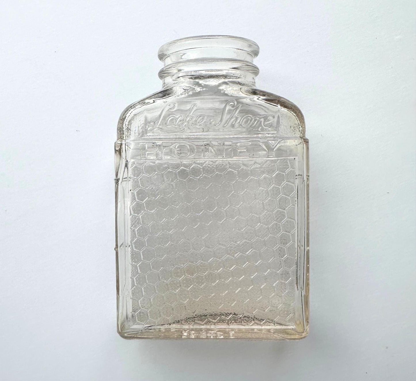 A vintage “Lake Shore HONEY” glass bottle shown in a flat-lay format against a soft white background. The image highlights the jar’s clear glass construction, hexagonal honeycomb embossing, and curved neck detail. A perfect visual for rustic fall decor or eco-conscious vintage collections, this 1950s bottle can be repurposed for dried florals, pantry storage, or minimalist shelf styling. The flat angle emphasizes its clean, geometric lines and sustainable design.