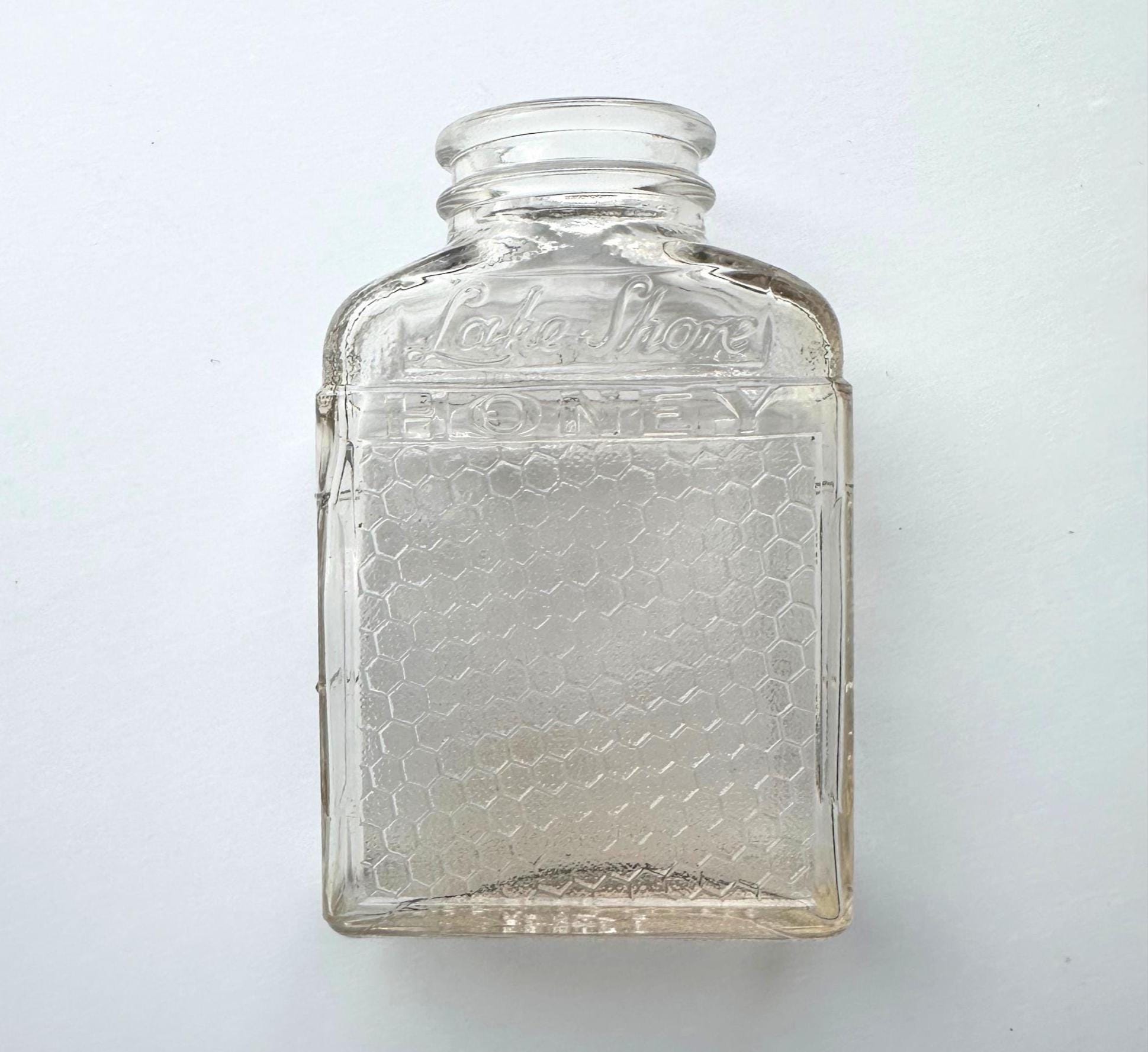 A vintage “Lake Shore HONEY” glass bottle shown in a flat-lay format against a soft white background. The image highlights the jar’s clear glass construction, hexagonal honeycomb embossing, and curved neck detail. A perfect visual for rustic fall decor or eco-conscious vintage collections, this 1950s bottle can be repurposed for dried florals, pantry storage, or minimalist shelf styling. The flat angle emphasizes its clean, geometric lines and sustainable design.