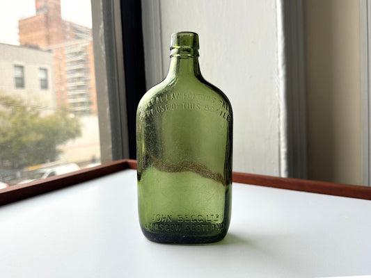 Hero image of the green John Begg Ltd. whiskey bottle showing both embossed front and curved shoulders. With moody fall light and New York cityscape in the background, this vintage glass piece fits perfectly into seasonal decor and reclaimed collections.