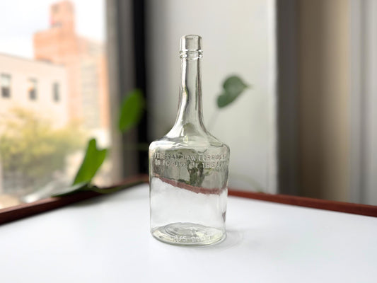 Vintage 4/5 quart clear glass liquor bottle 9.5 inches tall by 3.75 inches diameter, rescued from NYC beaches. Features embossed "FEDERAL LAW FORBIDS SALE OR RE-USE OF THIS BOTTLE" and "4/5 QUART" markings from the regulated spirits era 1935-1960s. Authentic mid-century American liquor bottle perfect for bar cart decor, vintage spirits collections, or tall single stem vases. Beach-rescued bottle with age-appropriate clarity.