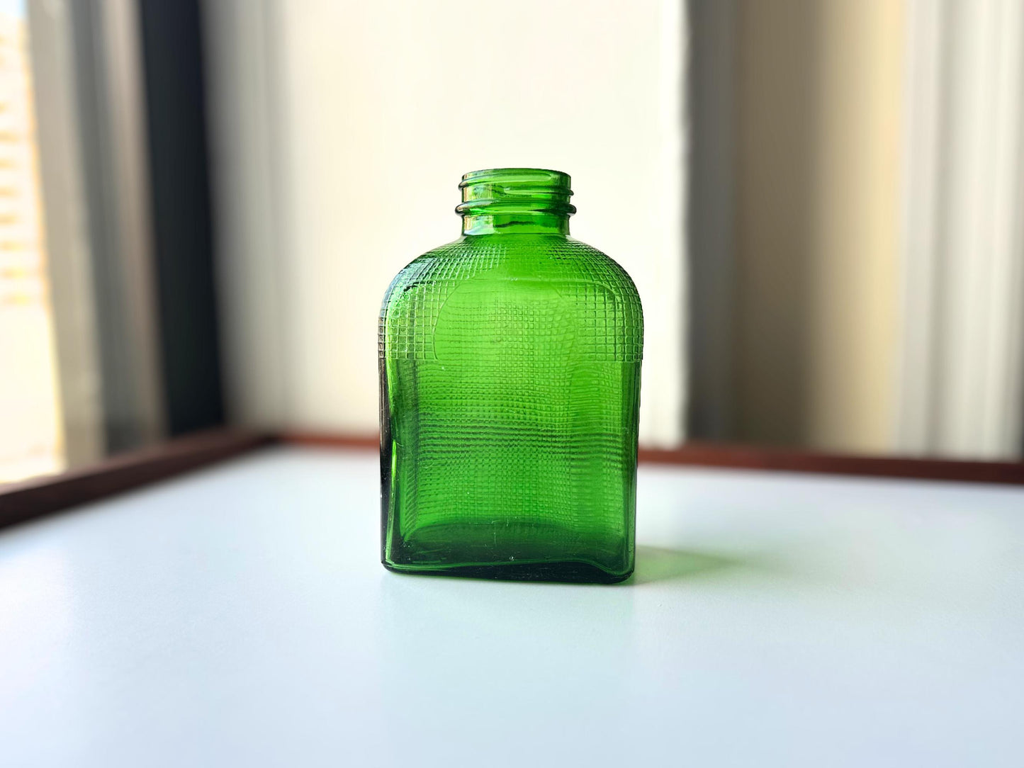 Stunning vintage emerald green glass bottle with distinctive waffle-grid texture throughout perfect for mid-century collectors farmhouse decor. Authentic 1950 bottle features tactile raised square pattern squared flask shape catches light beautifully. Rich emerald green three-dimensional textured surface. Ideal for windowsill displays vintage bottle collections retro kitchen shelves small bud vases statement pieces open shelving MCM cottagecore maximalist vintage interiors sustainable.