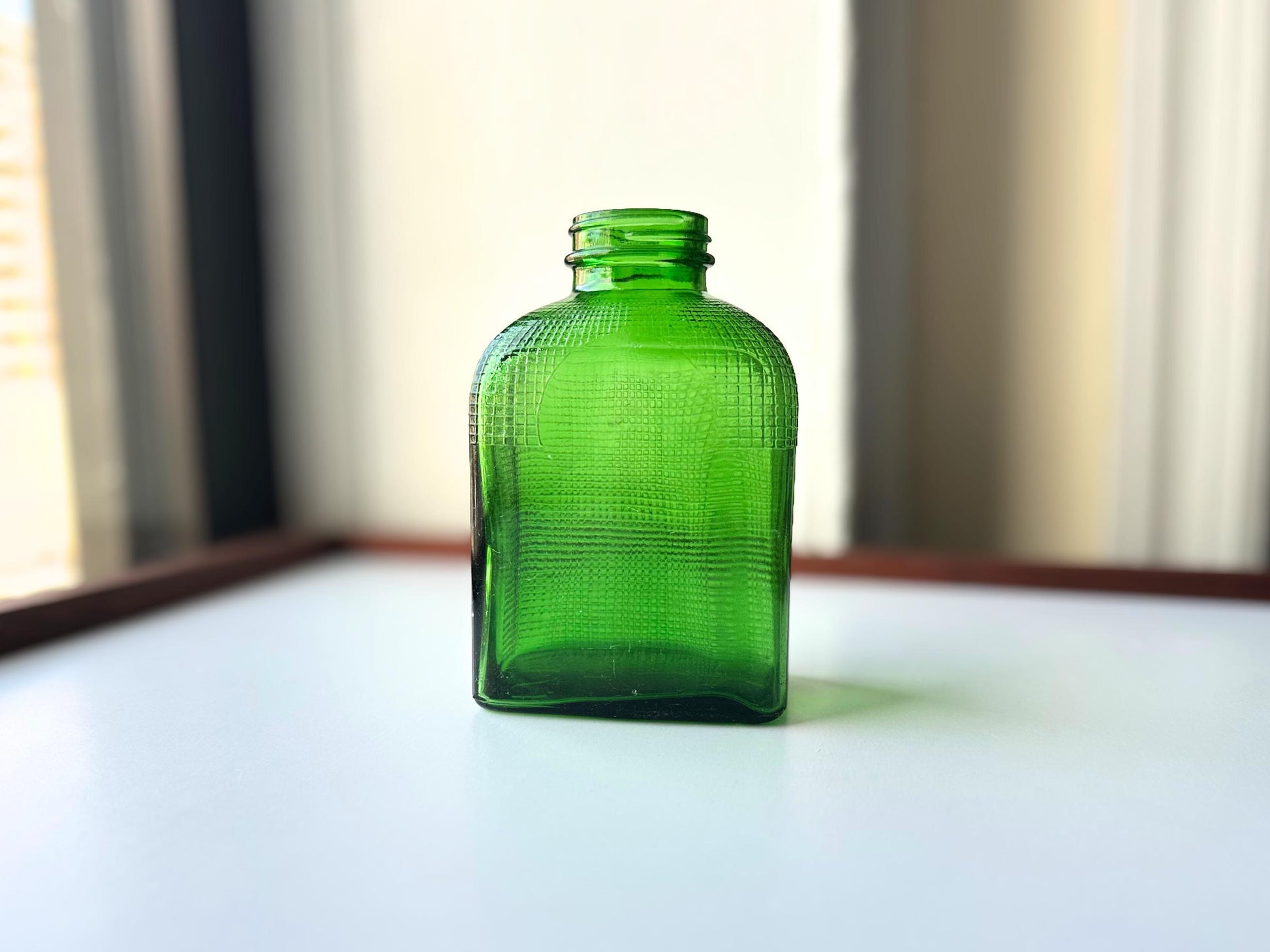 Stunning vintage emerald green glass bottle with distinctive waffle-grid texture throughout perfect for mid-century collectors farmhouse decor. Authentic 1950 bottle features tactile raised square pattern squared flask shape catches light beautifully. Rich emerald green three-dimensional textured surface. Ideal for windowsill displays vintage bottle collections retro kitchen shelves small bud vases statement pieces open shelving MCM cottagecore maximalist vintage interiors sustainable.