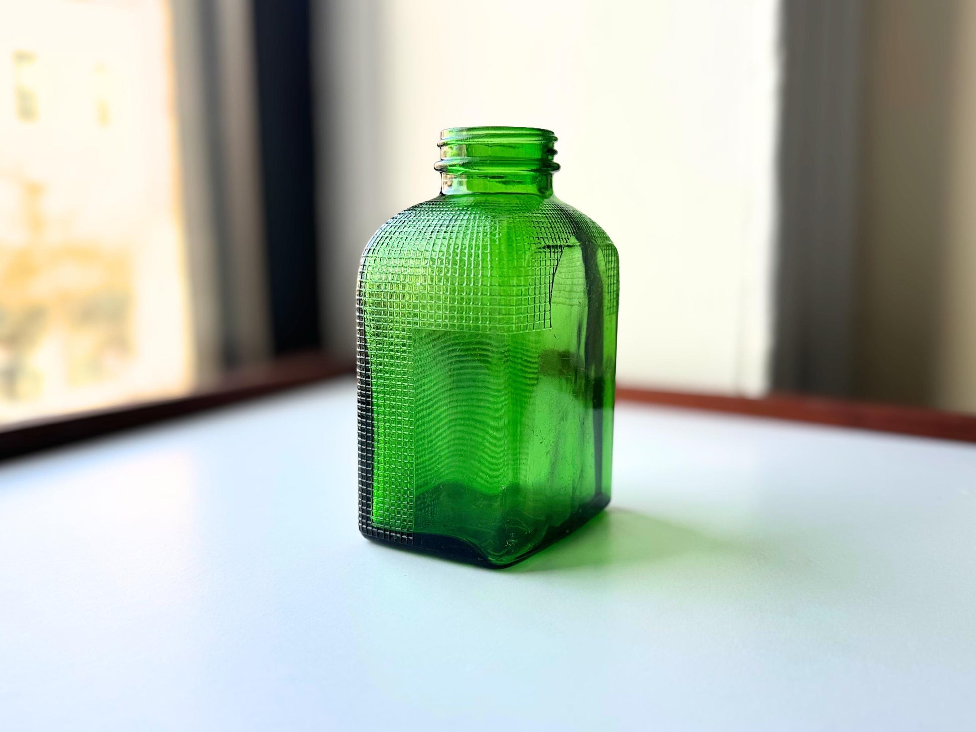 Stunning vintage emerald green glass bottle with distinctive waffle-grid texture throughout perfect for mid-century collectors farmhouse decor. Authentic 1950 bottle features tactile raised square pattern squared flask shape catches light beautifully. Rich emerald green three-dimensional textured surface. Ideal for windowsill displays vintage bottle collections retro kitchen shelves small bud vases statement pieces open shelving MCM cottagecore maximalist vintage interiors sustainable.