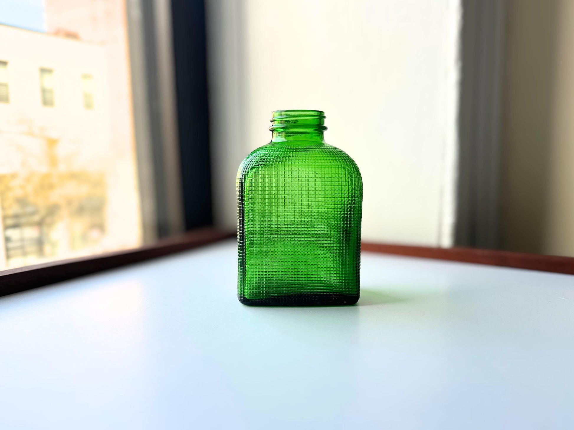 Stunning vintage emerald green glass bottle with distinctive waffle-grid texture throughout perfect for mid-century collectors farmhouse decor. Authentic 1950 bottle features tactile raised square pattern squared flask shape catches light beautifully. Rich emerald green three-dimensional textured surface. Ideal for windowsill displays vintage bottle collections retro kitchen shelves small bud vases statement pieces open shelving MCM cottagecore maximalist vintage interiors sustainable.