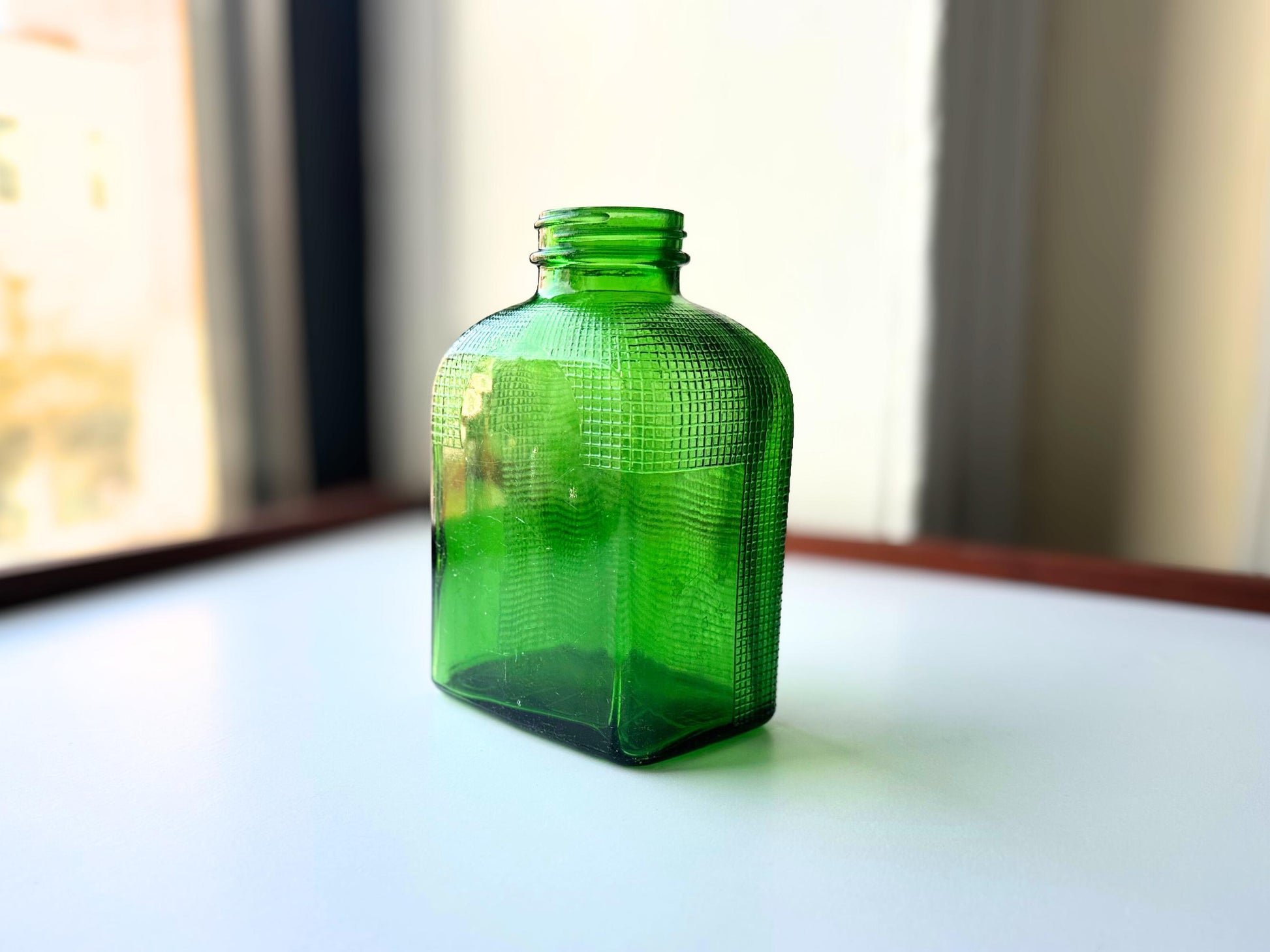 Stunning vintage emerald green glass bottle with distinctive waffle-grid texture throughout perfect for mid-century collectors farmhouse decor. Authentic 1950 bottle features tactile raised square pattern squared flask shape catches light beautifully. Rich emerald green three-dimensional textured surface. Ideal for windowsill displays vintage bottle collections retro kitchen shelves small bud vases statement pieces open shelving MCM cottagecore maximalist vintage interiors sustainable.