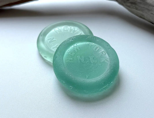 Two round, translucent sea glass vintage coca cola bottoms in a light mint green color, resting on a white surface next to a piece of natural driftwood.