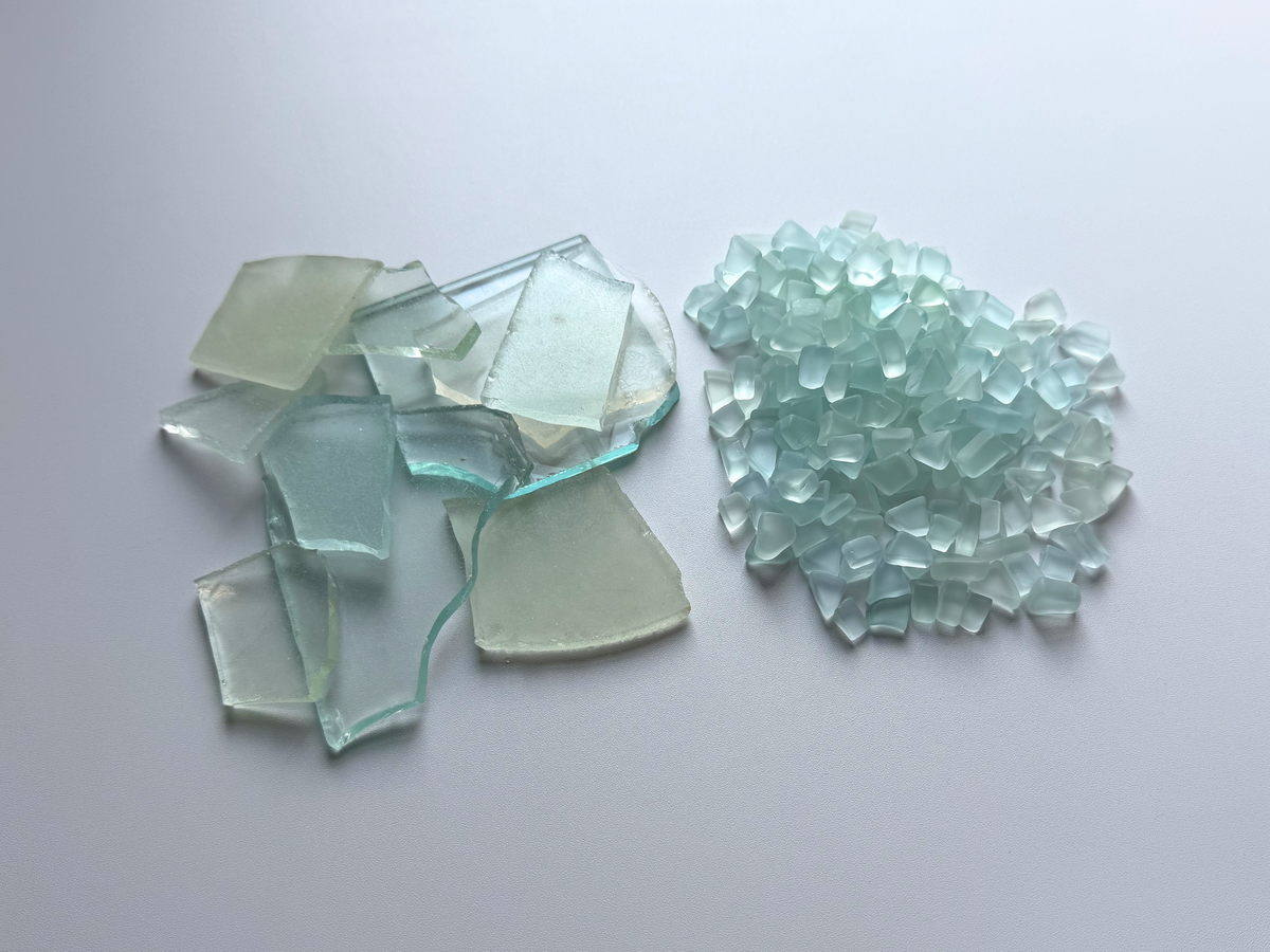 Side-by-side photo showing a raw glass shard held in a gloved hand at an NYC beach cleanup site next to a pile of finished pale seafoam and sky blue tumbled sea glass, illustrating how 100% beach-found household glass is recovered and tumbled in-house to a smooth finish.