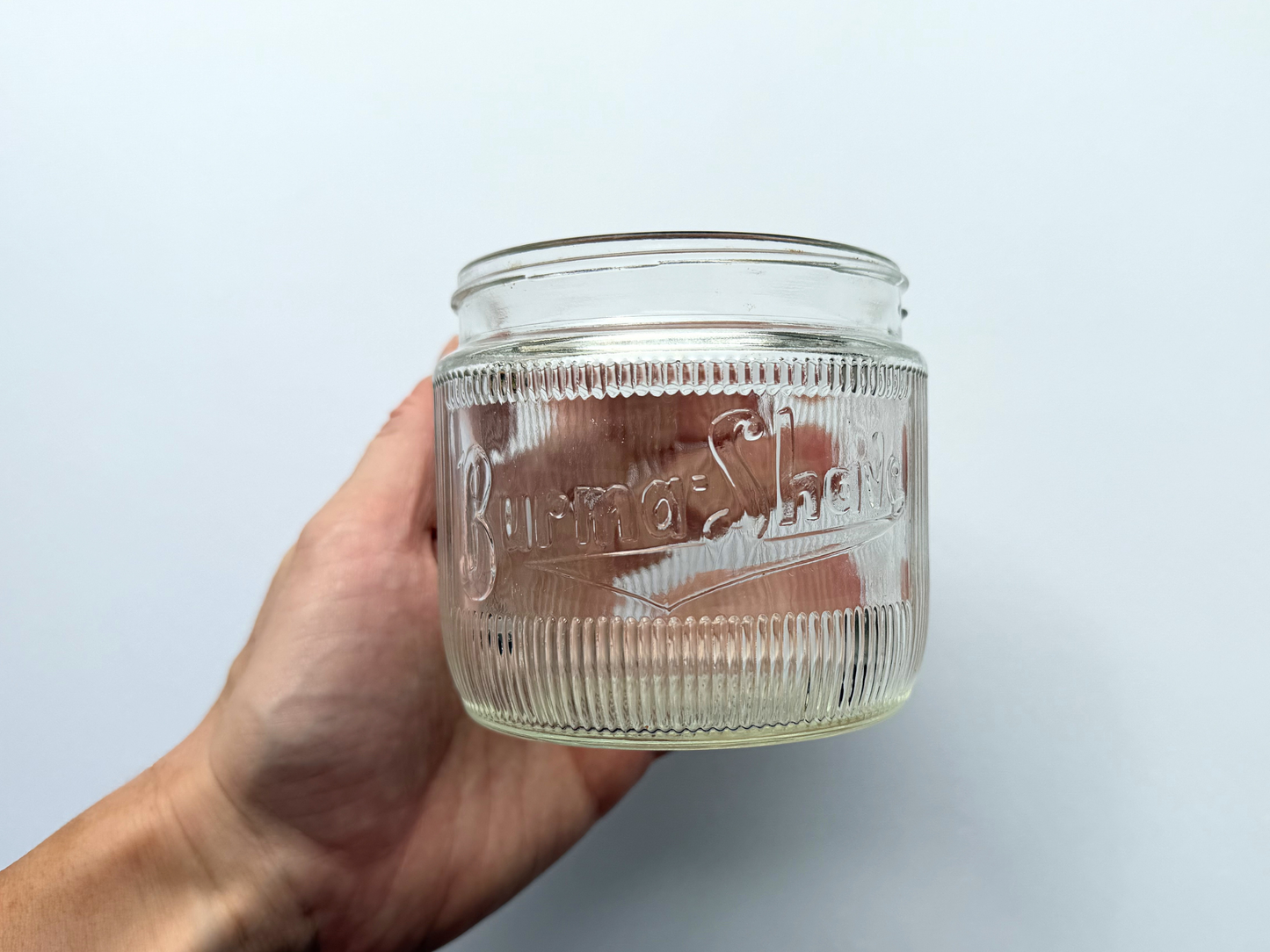 A hand holding a vintage ribbed glass Burma-Shave jar, providing a scale reference that shows its compact 3.4-inch height.