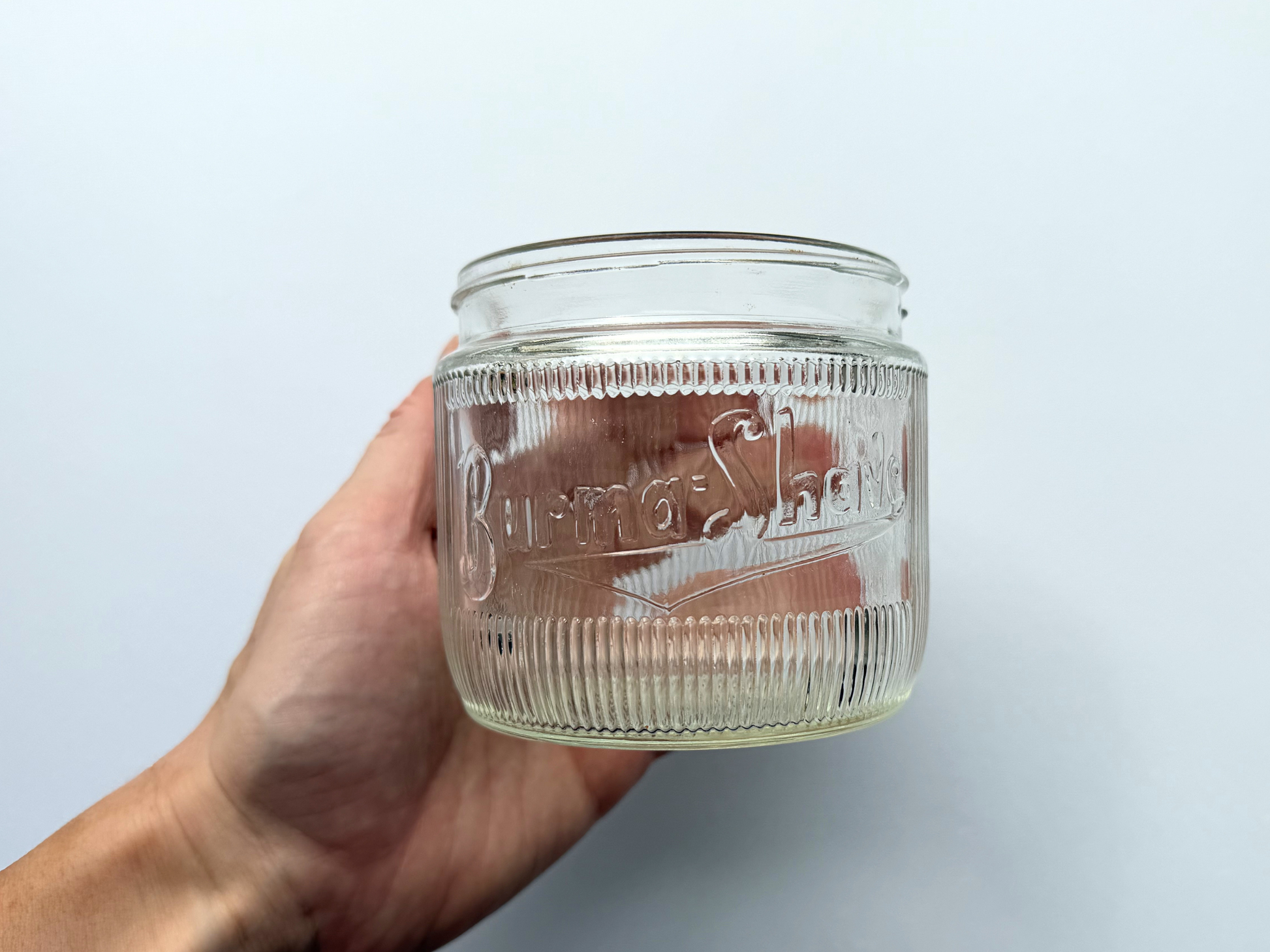 A hand holding a vintage ribbed glass Burma-Shave jar, providing a scale reference that shows its compact 3.4-inch height.