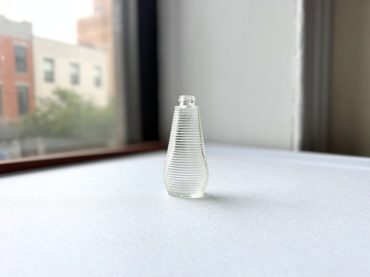 Small vintage clear glass ribbed bottle with a rounded halo shoulder, once used for shampoo, shown at a three-quarter angle highlighting its textured surface.