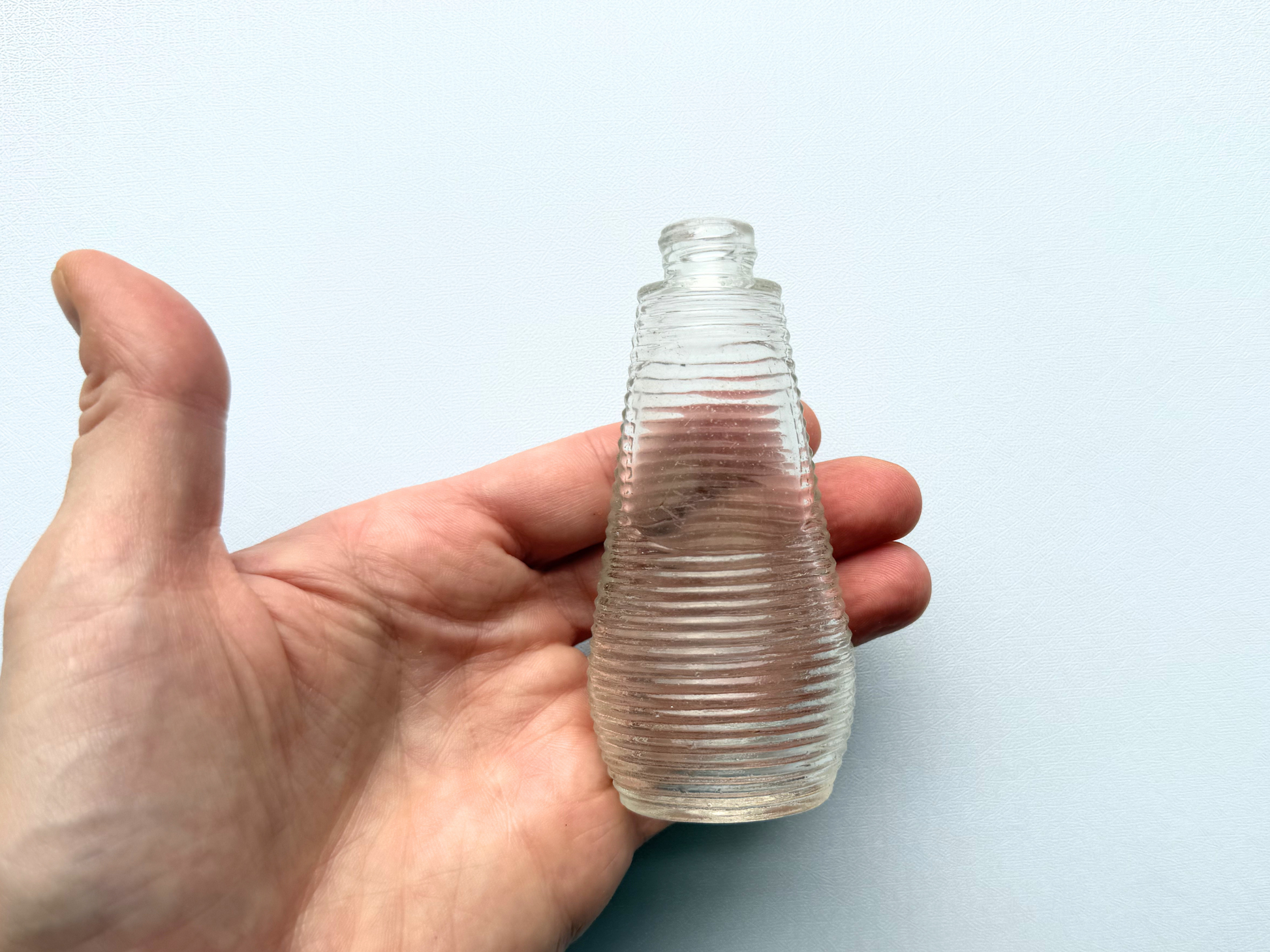 A hand holding a small vintage ribbed glass halo bottle, providing a scale reference that shows its miniature 3.85-inch height.