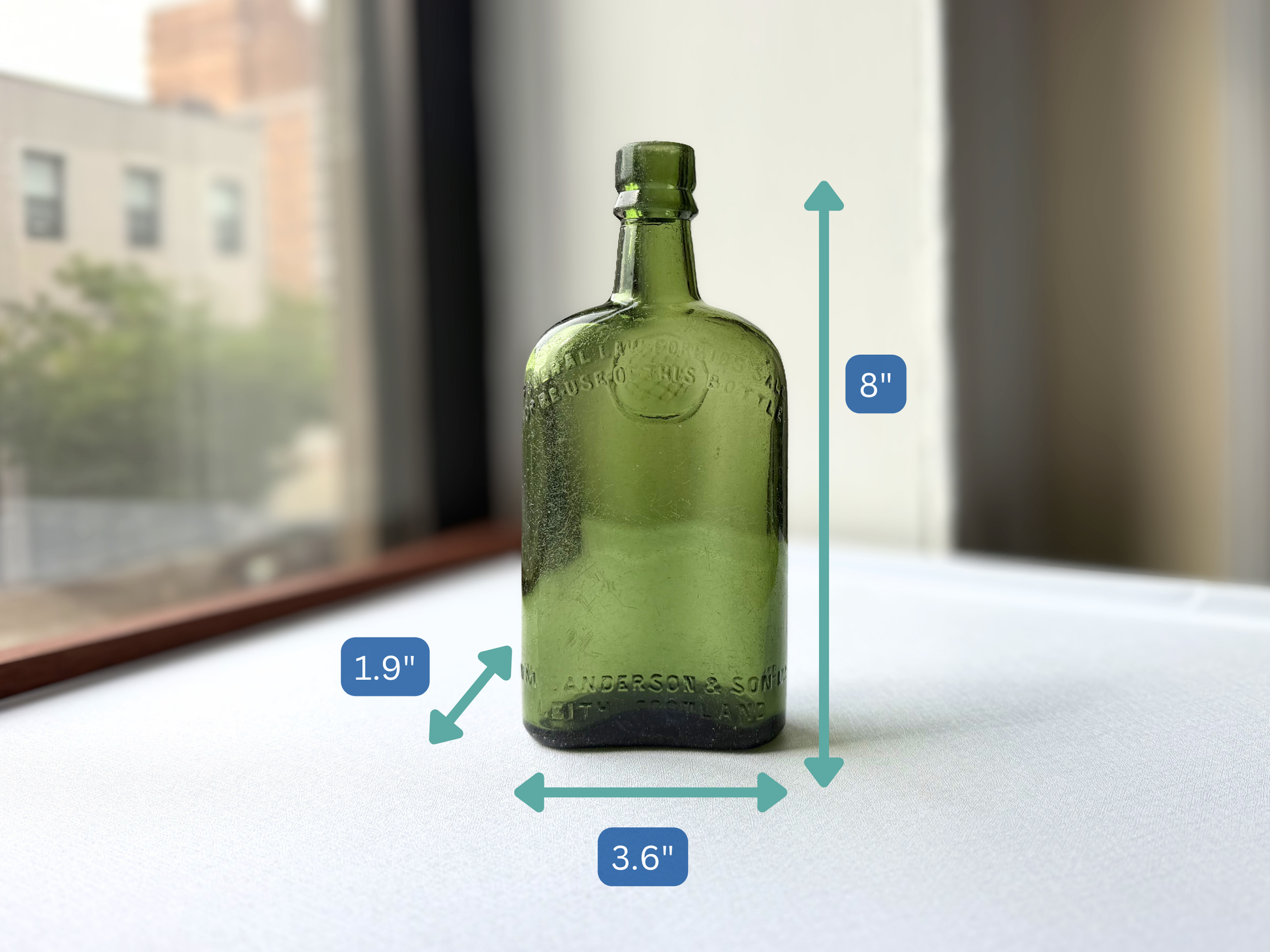Vintage green glass Sanderson bottle shown with measurement markers indicating a height of 8 inches, width of 3.6 inches, and depth of 1.9 inches, illustrating its substantial scale for shelf or bar cart display.