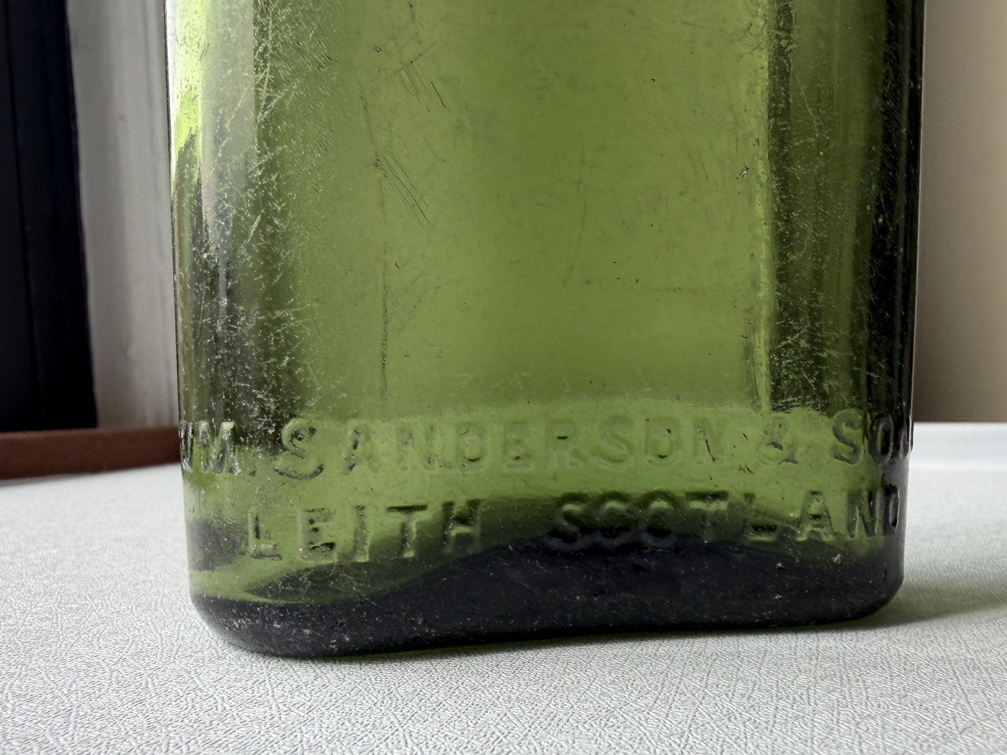 Close-up of the embossed lettering reading William Sanderson & Son Ltd. Leith Scotland on the front of a vintage green glass bottle, with sharp, legible text characteristic of quality mid-century glasswork.