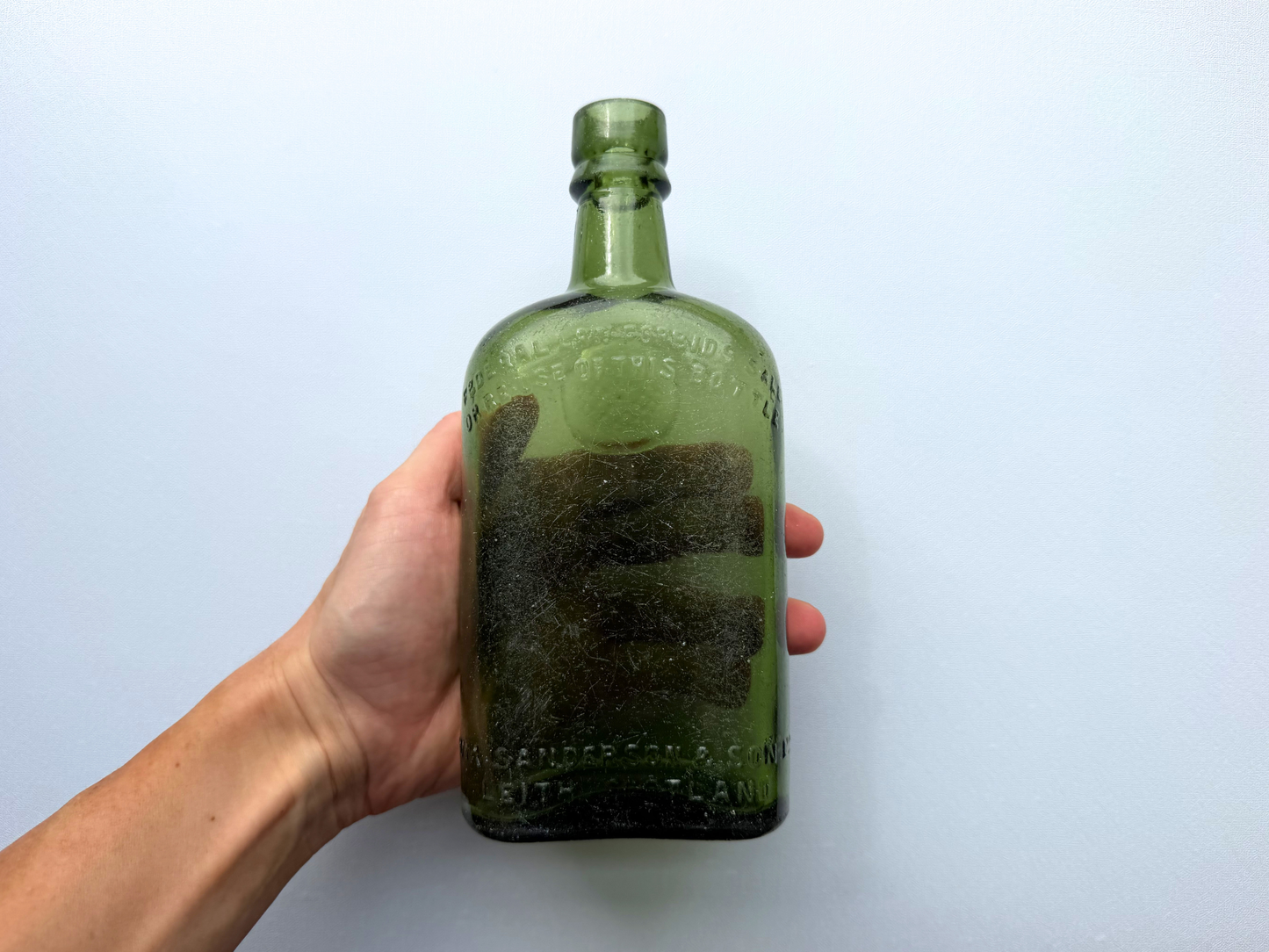 A hand holding a vintage green glass William Sanderson bottle, providing a scale reference that shows its substantial 8-inch height and rounded, weighty form.
