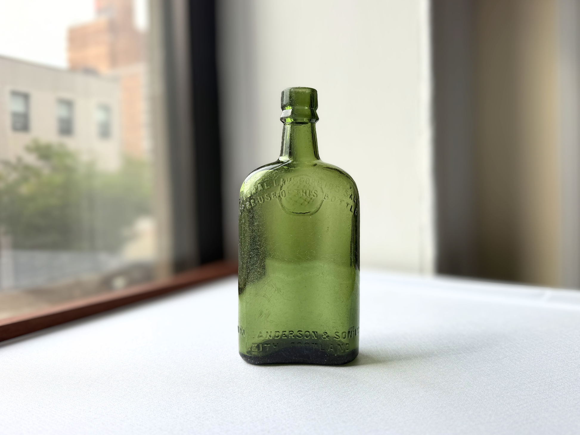 Side angle view of a vintage green glass William Sanderson bottle, highlighting its rounded profile and the subtle curvature that makes it a striking Neo Deco style accent.